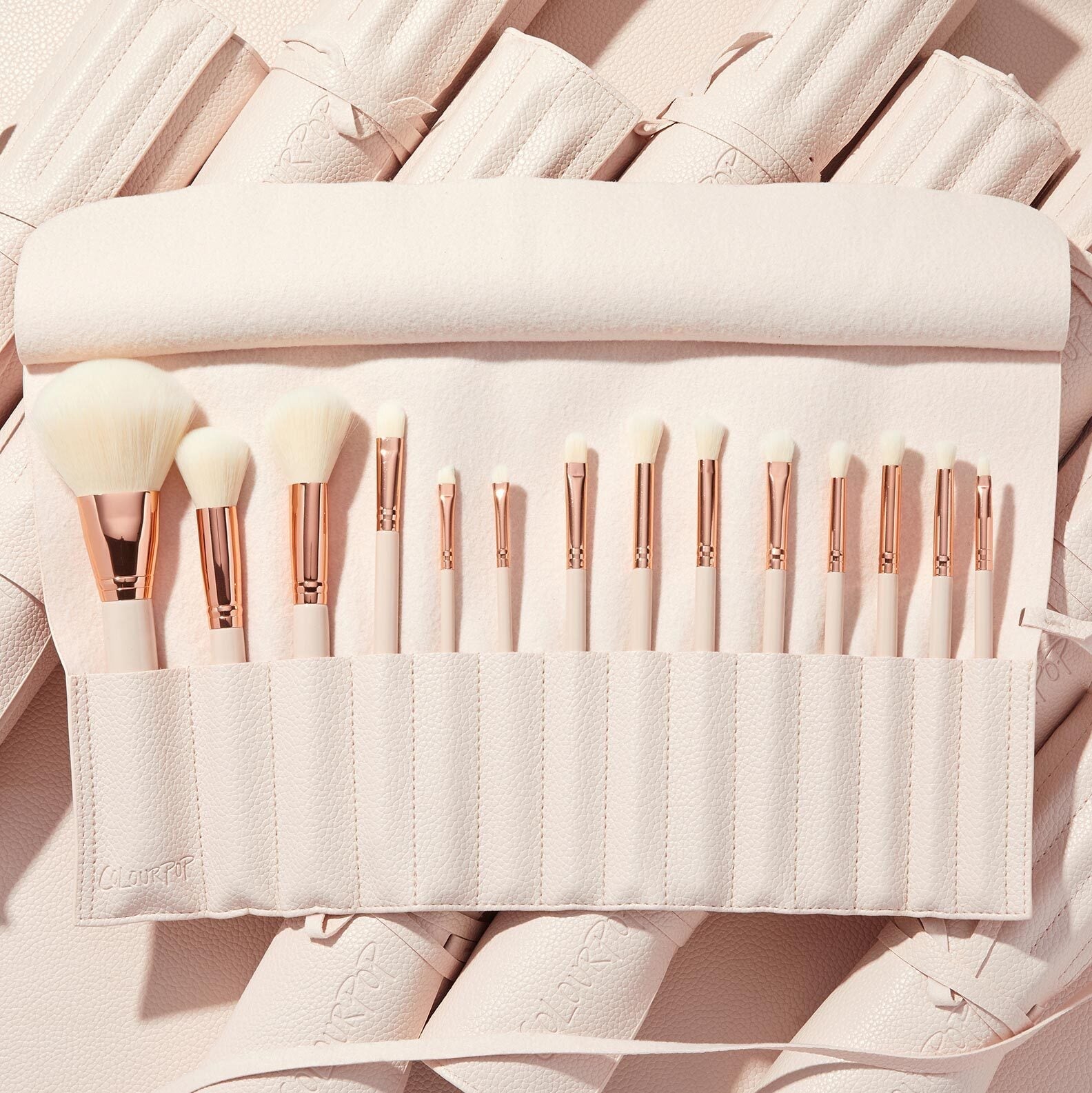 Ultimate Brush Roll displaying a full set of soft white-bristle makeup brushes with rose-gold ferrules neatly slotted in a blush pink travel roll, set on matching rolled cases - ColourPop Makeup Brush Kit for face and eye application.