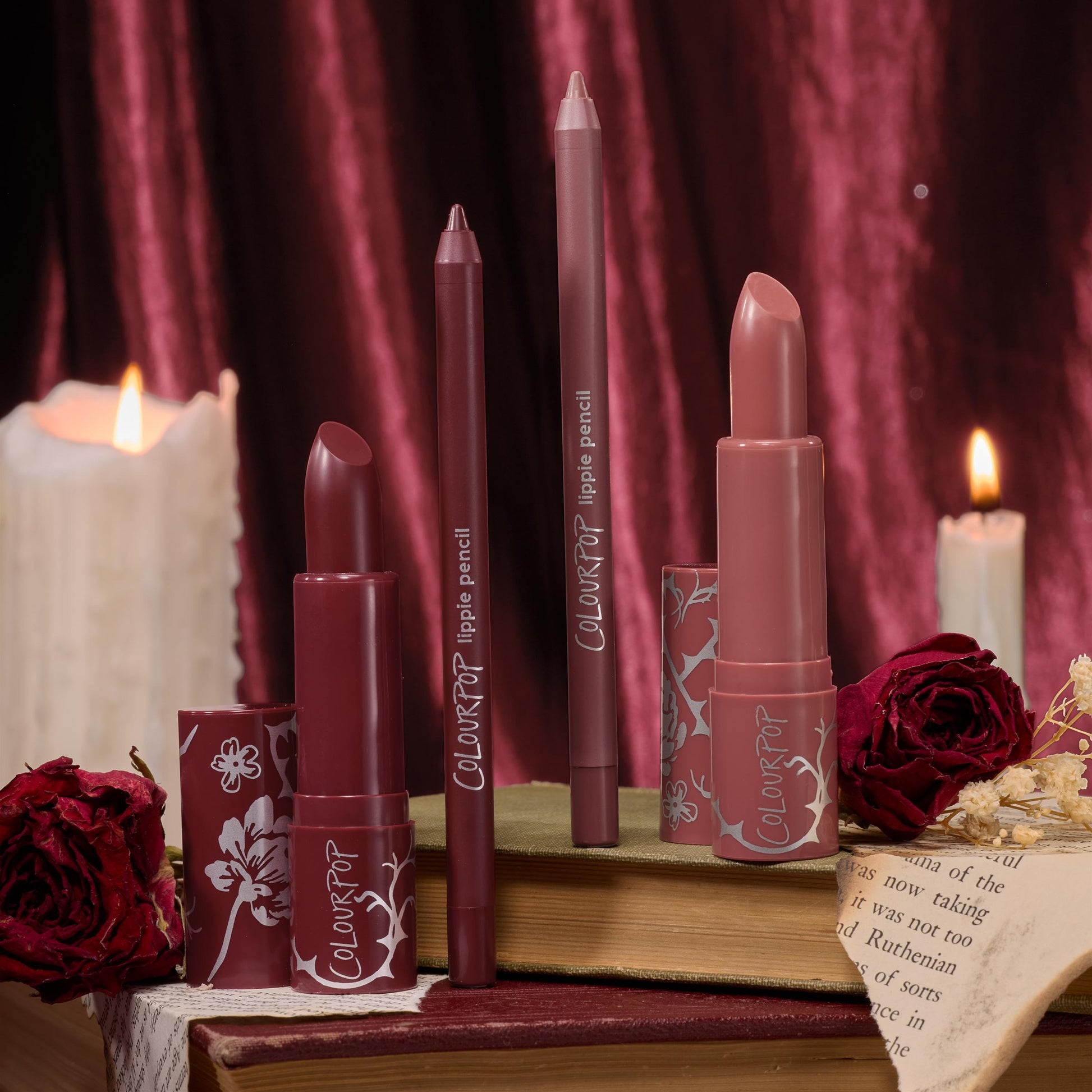 Secret Kiss ColourPop Lipstick & Lip Pencil Set - two creamy lipsticks and two matching lippie pencils in deep berry and rosy nude - standing upright in floral-print tubes on vintage books amid roses and lit candles, burgundy velvet backdrop.