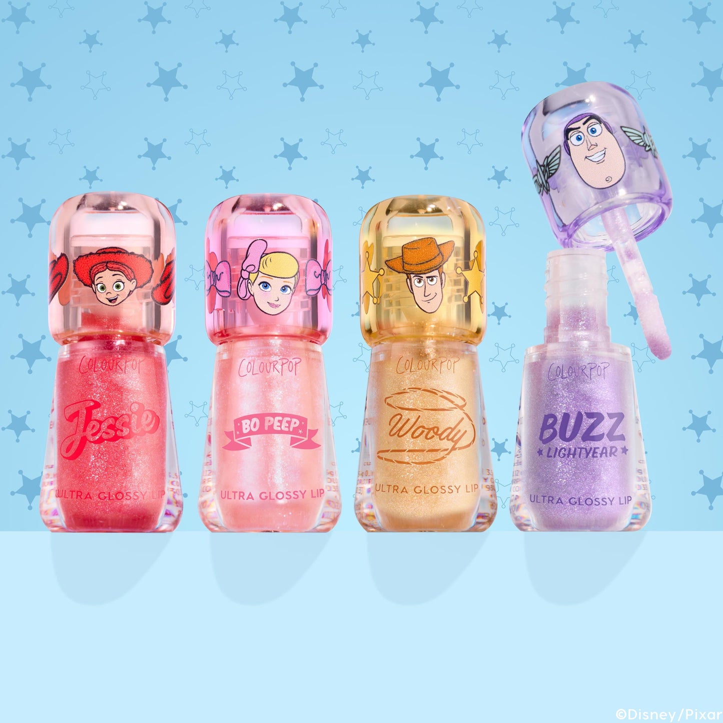 Squad Dreams ColourPop Ultra Glossy Lip bundle lined up: Jessie coral pink shimmer, Bo Peep soft pink shimmer, Woody warm gold glitter, Buzz Lightyear lavender sparkle with wand lifted; character caps; light blue Toy Story sheriff-star backdrop.