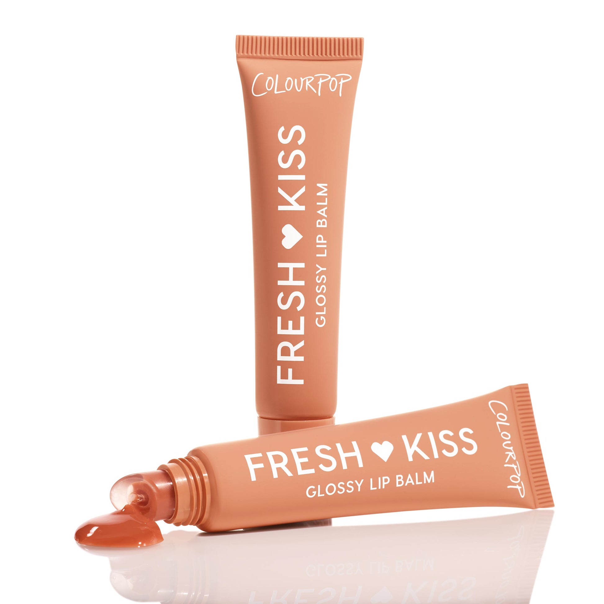 ColourPop Fresh Kiss Glossy Balm in Caramel Latte, one tube standing while another uncapped squeezes out a caramel-brown gloss droplet on a white reflective surface, showcasing the warm nude shade and soft-touch squeeze-tube packaging.