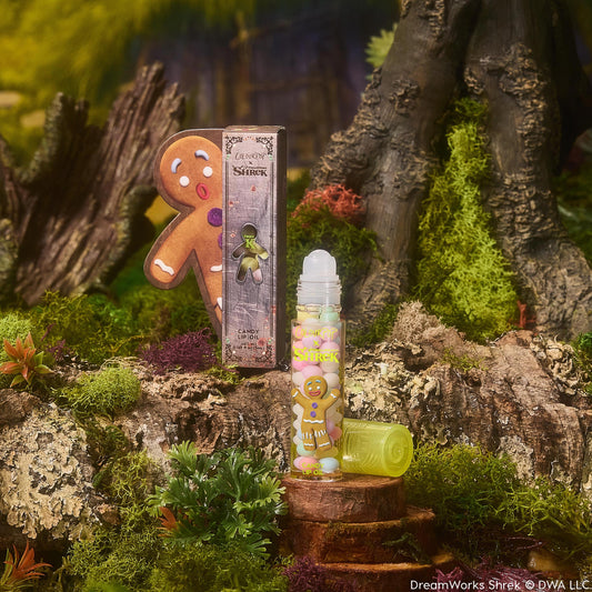 Gumdrop Buttons ColourPop Candy Lip Oil stands uncapped on a mossy stump, clear roll-on tube with gingerbread man graphic and lime cap beside it, posed with Shrek-themed packaging in a swampy forest setting.